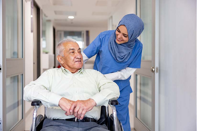 A nurse in blue scrubs and a hijab is gently pushing an elderly gentleman in a wheelchair through a hospital corridor. The nurse is leaning forward slightly, smiling and engaging with the patient, who looks relaxed and content.