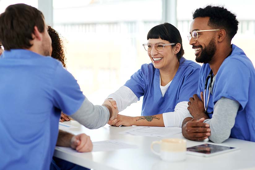A group of nurses wearing blue scrubs are seated around a conference table and smiling. Two nurses are shaking hands across the table.