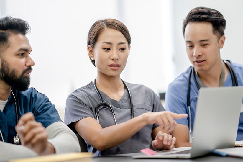 A diverse group of three (3) nursing students working together on a group project. The female nursing student is seated in the middle and is pointing at the laptop screen while talking with her male classmates.