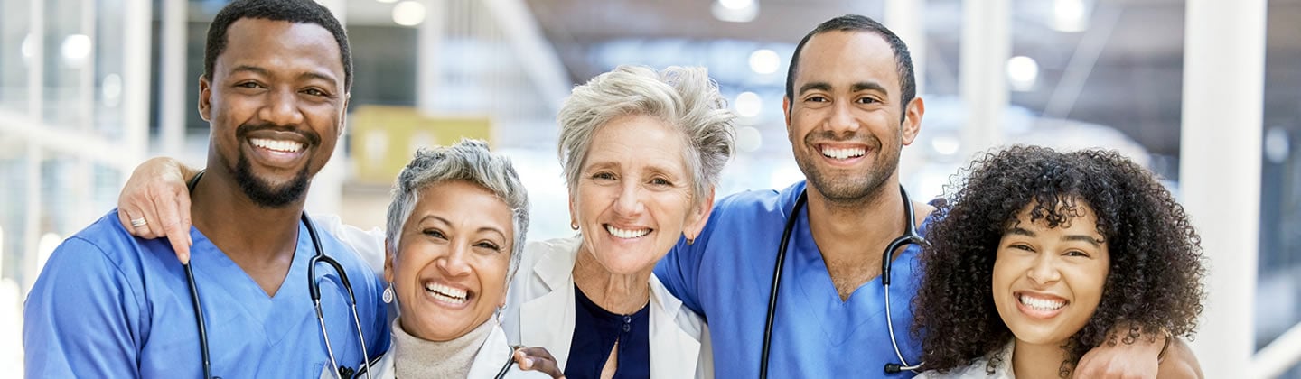 Diverse group of healthcare professionals smiling together, wearing scrubs and white coats, with stethoscopes around their necks.