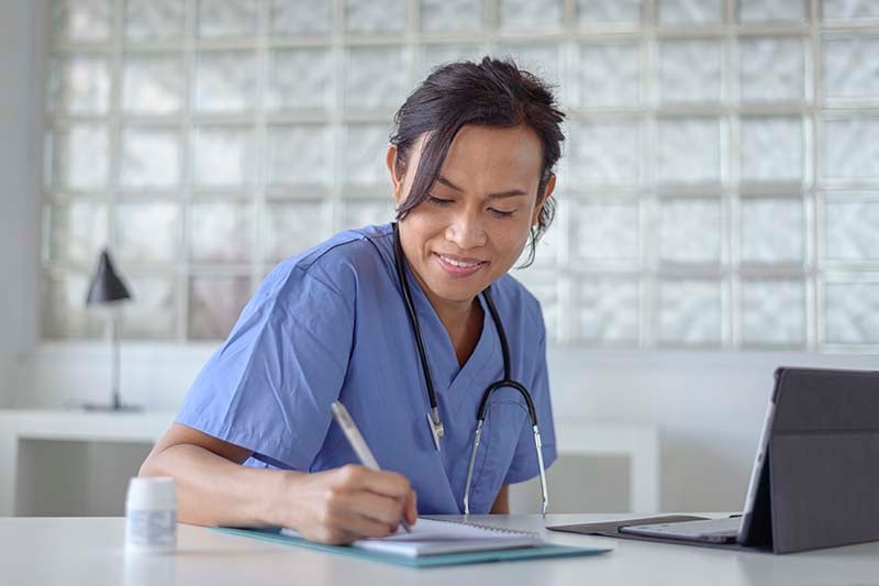 A young female nurse is seated at a desk, wearing a light blue scrub outfit and doing research using a laptop and taking notes.