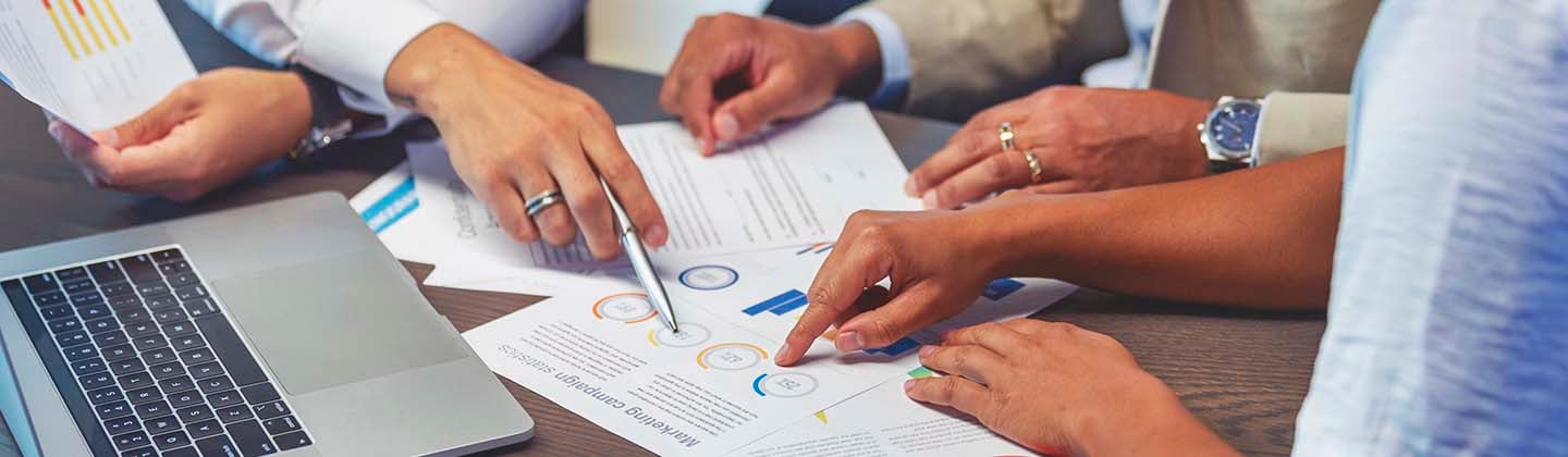 Three people are seated at a table and reviewing financial data spread out before them. Only their arms and hands are visible as they point to sections of a graphic. 