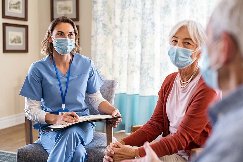 A nurse wearing a mask and sitting in a chair with a person in a room with a table