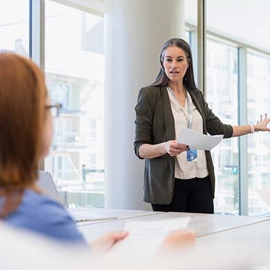 A female Director of Nursing stands in front of a conference table and is looking at a group of seated employees while giving a presentation and pointing to some information on a whiteboard behind her.