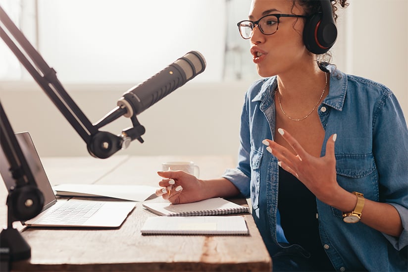 An Image of a female dressed in a denim shirt over a black t-shirt. She is seated at a desk, and she is wearing headphones. There is a professional microphone on a stand in front of her, and she is speaking into the microphone and gesturing with her hands to show she is conveying a message.