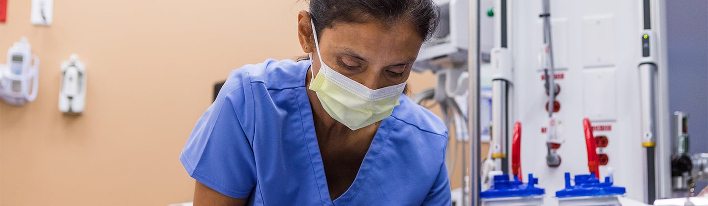 A caring emergency room nurse in blue scrubs is leaning over a patient and stroking their hair while adjusting their oxygen mask. The patient is calmy lying on a stretcher with their eyes closed.