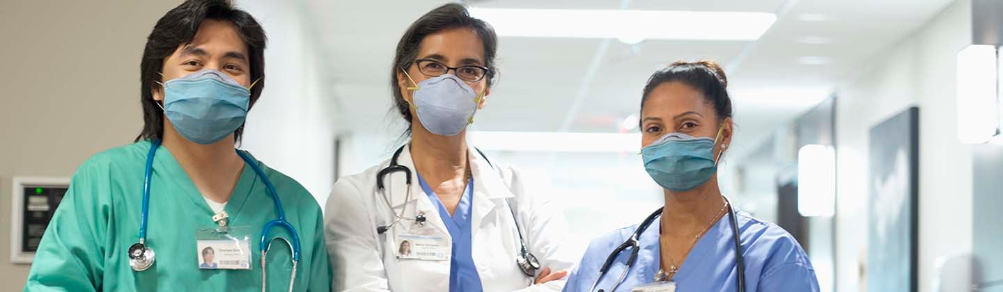 A diverse team of nurses gathers around a table for a meeting, with one standing and leading the discussion, illustrating collaboration and engagement in a clinical setting. 