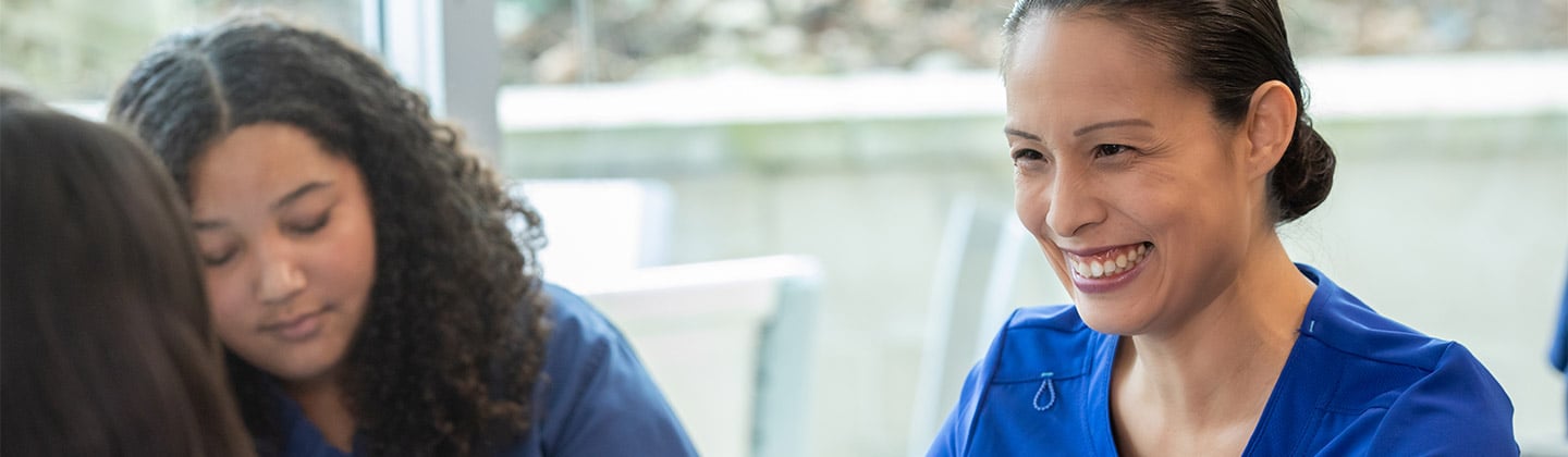 This image captures a cheerful nurse coach in blue scrubs engaging with a student or colleague across a table. She is smiling, gesturing with her hands, illustrating a point or giving advice. A laptop is open in front of her, suggesting a learning or mentorship session. The focus on her face and the attentive gaze of the young woman listening reflects a supportive educational interaction in a bright, welcoming setting.