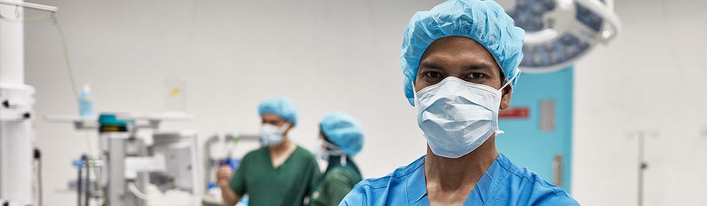 A confident nurse in blue scrubs and a surgical cap stands with arms crossed in an operating room, with medical equipment and colleagues working in the background.