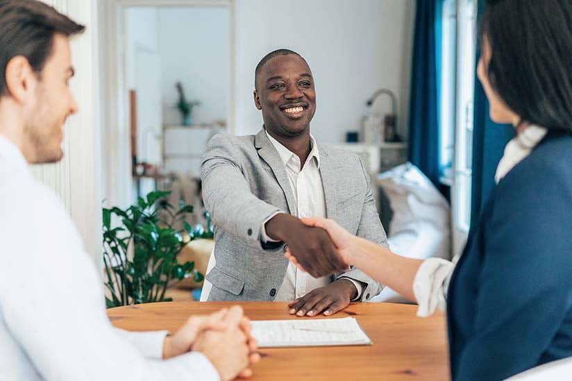 A confident and smiling male nurse is seated across a table from two interviewers, and he is reaching forward and shaking hands.