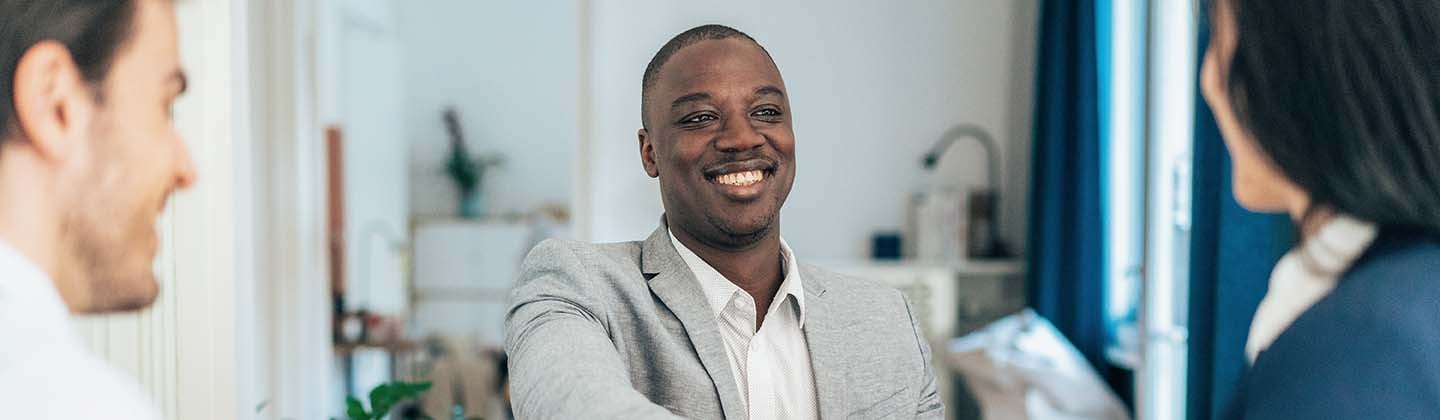 A confident and smiling male nurse is seated across a table from two interviewers, and he is reaching forward and shaking hands.