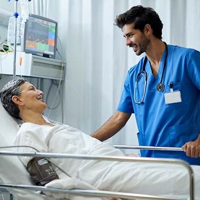 A%20male%20nurse%20is%20standing%20at%20the%20bedside%20of%20his%20hospitalized%20patient,%20smiling,%20and%20looking%20attentively.%20The%20patient%20is%20reclining%20and%20is%20attached%20to%20a%20blood%20pressure%20monitor%20and%20a%20nasal%20cannula.