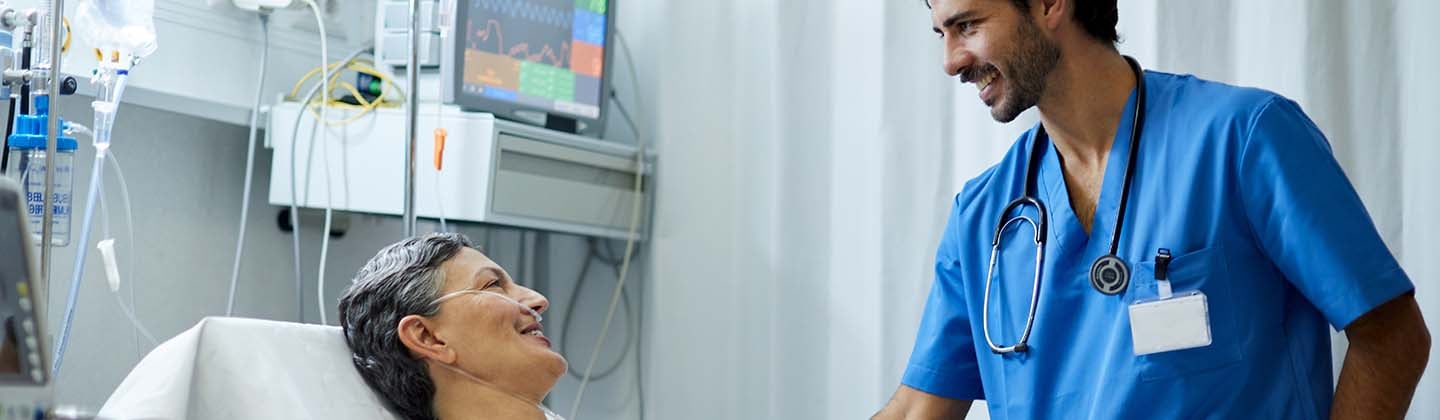 A male nurse is standing at the bedside of his hospitalized patient, smiling, and looking attentively. The patient is reclining and is attached to a blood pressure monitor and a nasal cannula.