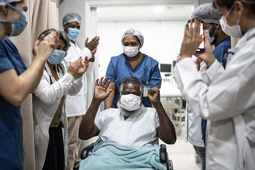 Elderly male patient is being wheeled out of the hospital and hospital staff have lined the hallway and are clapping in happiness for his recovery.