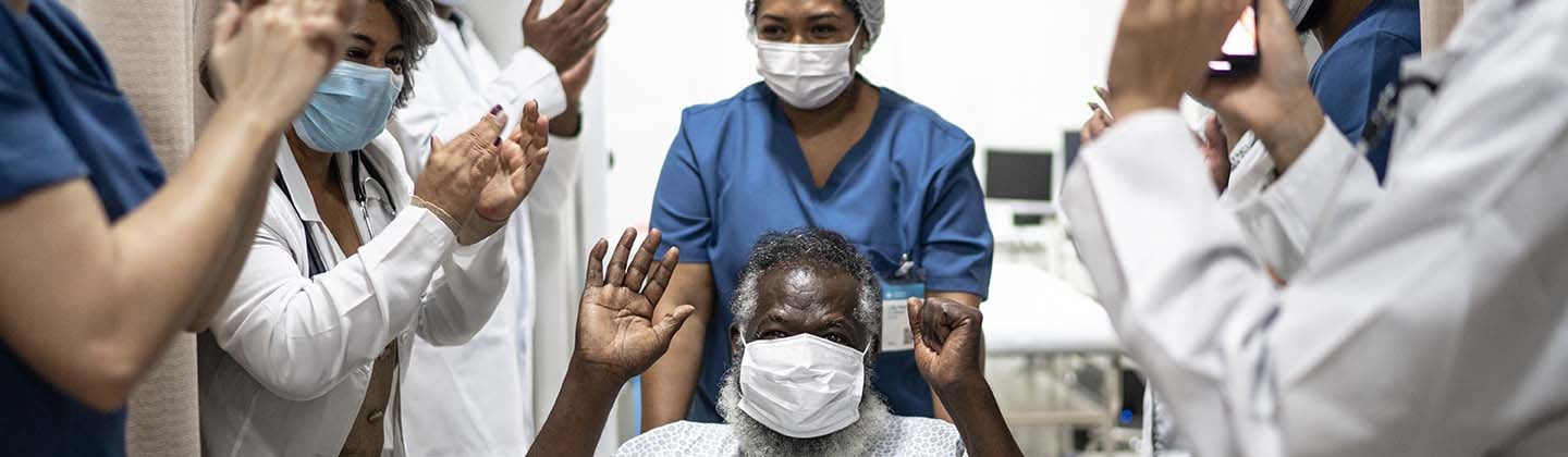 Elderly male patient is being wheeled out of the hospital and hospital staff have lined the hallway and are clapping in happiness for his recovery.