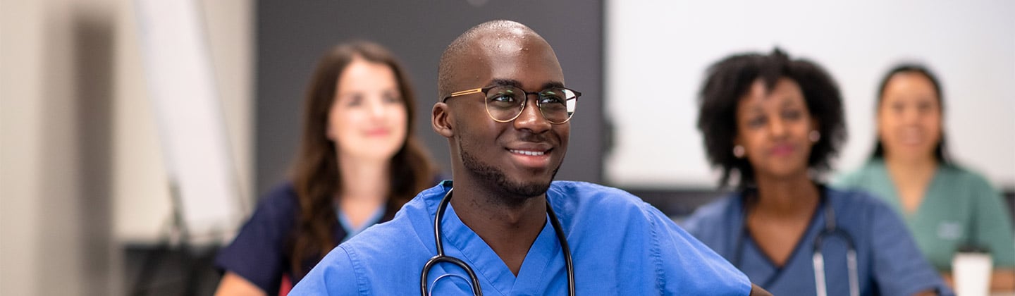 A diverse group of nursing students dressed in various color scrubs are engaged in learning in a classroom setting.