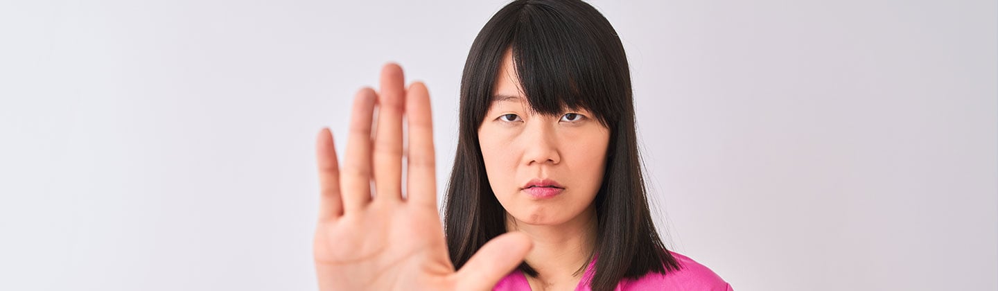 A female nurse wearing a pink scrub top and a stethoscope around her neck is in front of a white background and doing stop sign gesture with the palm of her hand. She has a serious expression on her face.