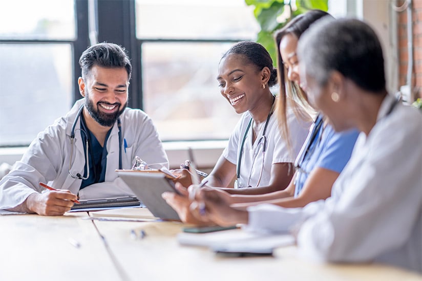 A small group of four medical professionals sit around a boardroom table as they meet to discuss patient cases. They are each dressed professionally in scrubs and lab coats as they focus on working together.