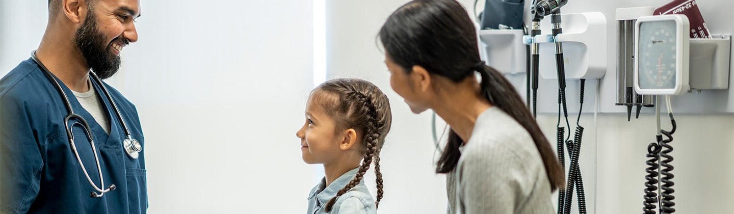 A cute seven year old girl is at a check-up appointment with her healthcare provider. She is sitting on the examination table with her mother. She is looking at her male nurse practitioner and smiling while her mother is seated next to her.