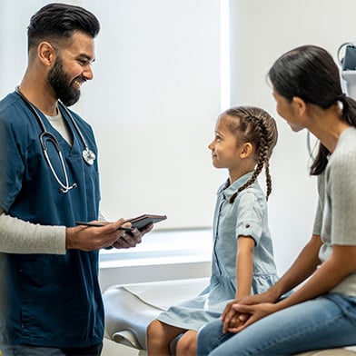 A cute seven year old girl is at a check-up appointment with her healthcare provider. She is sitting on the examination table with her mother. She is looking at her male nurse practitioner and smiling while her mother is seated next to her.