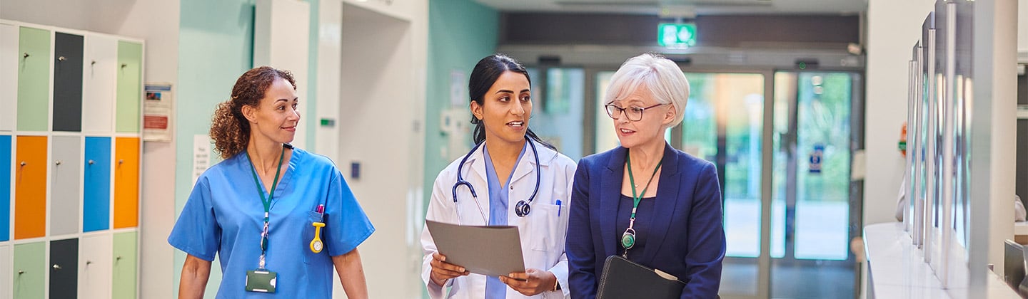 Female utilization nurse walking in a hospital hallway discussing a patient’s care with a female nurse and a female doctor.