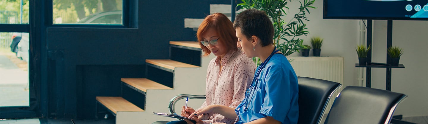 Two nurses, one in scrubs, discuss a patient's chart earnestly, embodying the principle of veracity in nursing through honest communication in a clinic waiting area.