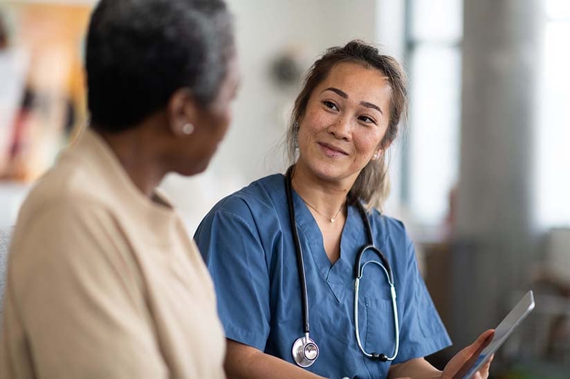 A cheerful nurse in blue scrubs takes notes on a clipboard while engaging with an elderly patient in a clinic room, signifying a caring and professional nurse-patient interaction.