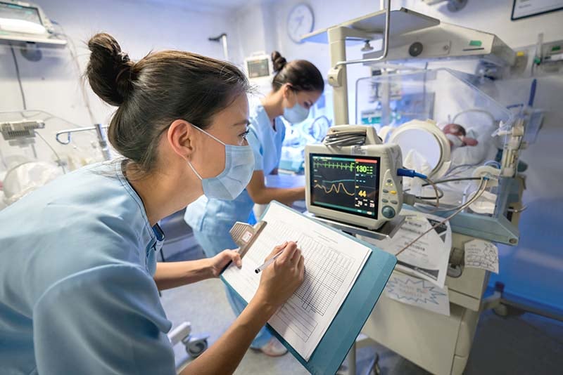 A female nurse is working in the Neo-Natal Intensive Care Unit. She is at the foot of an incubator documenting information from a monitoring device while a co-worker is checking on the baby.