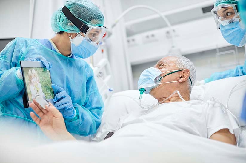 Two nurses are wearing isolation gowns, goggles, and facemasks, and are standing at either side of a patient’s bed. An elderly male patient is in the bed. He is also wearing a mask, and he is looking at a tablet screen the nurse is holding so he can talk with his family over the internet.