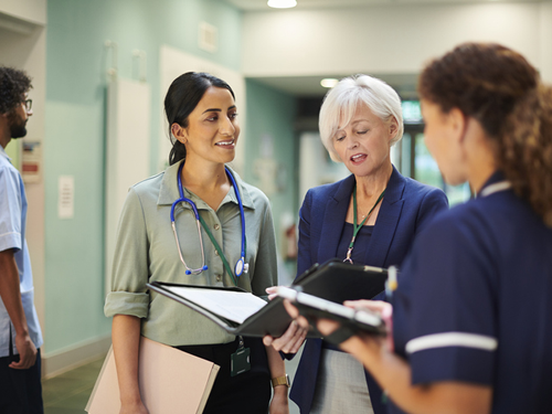 A group of nursing professionals are standing in a hallway of a medical center, and they are looking at a folder of data and discussing safety protocols.