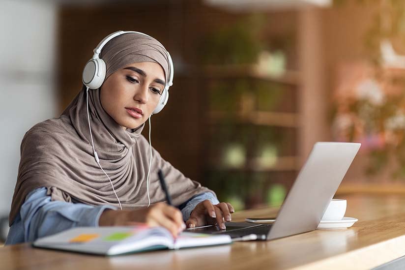 A focused nursing student in a hijab is studying for the NCLEX exam at a desk. She is wearing headphones and looking intently at her laptop screen while taking notes. A coffee cup and various study materials are organized around her, suggesting a productive study session in a quiet, indoor environment.