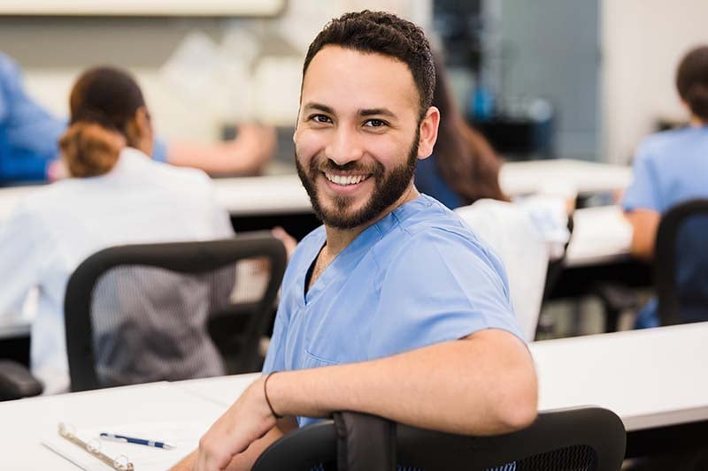 A smiling nurse in blue scrubs, seated at a table with medical equipment, looks back over his shoulder, representing a positive and dedicated healthcare learning environment.