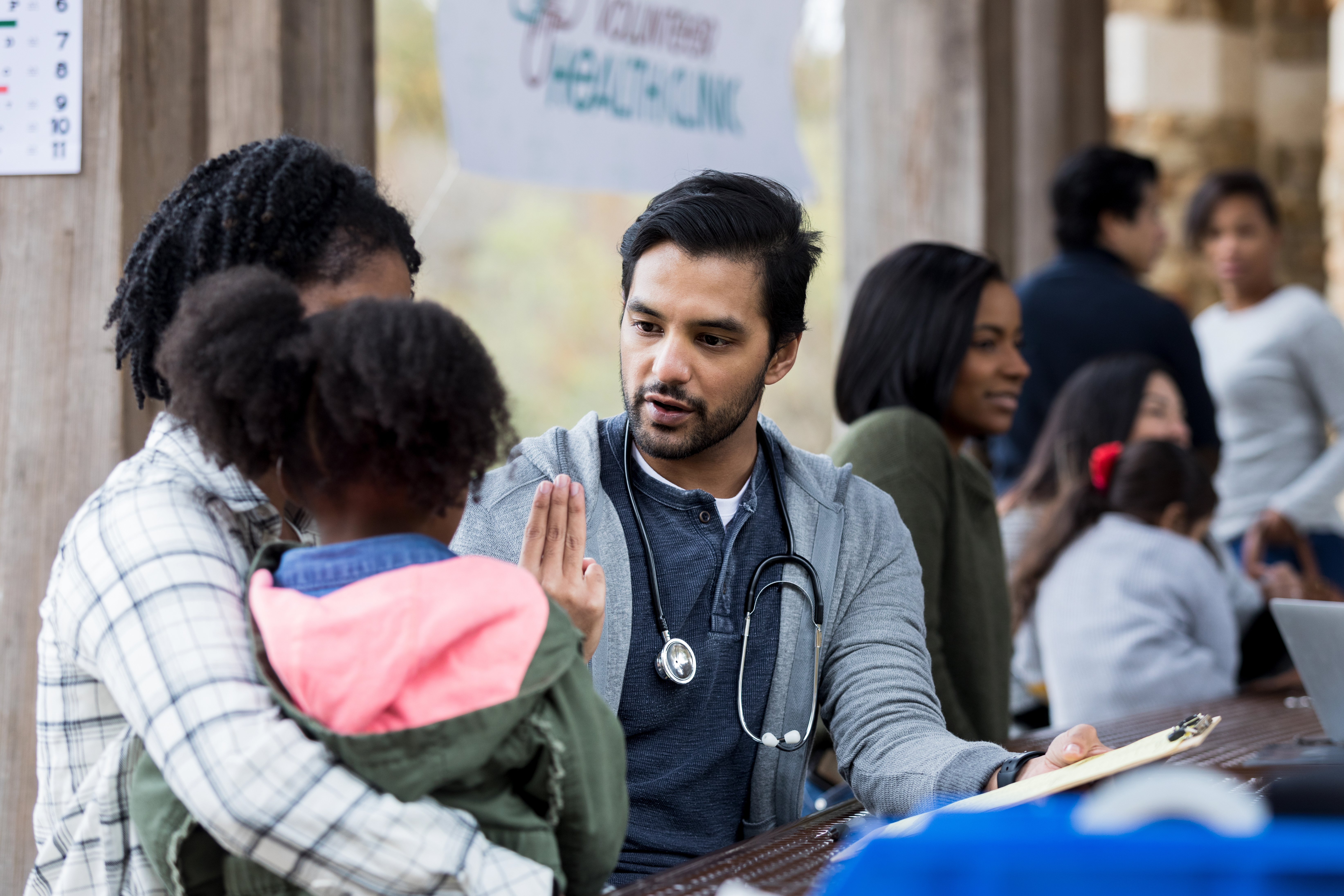 A nurse with a stethoscope engages with a child and her mother at a community health event, offering care and advice in an outdoor setting with other participants around.