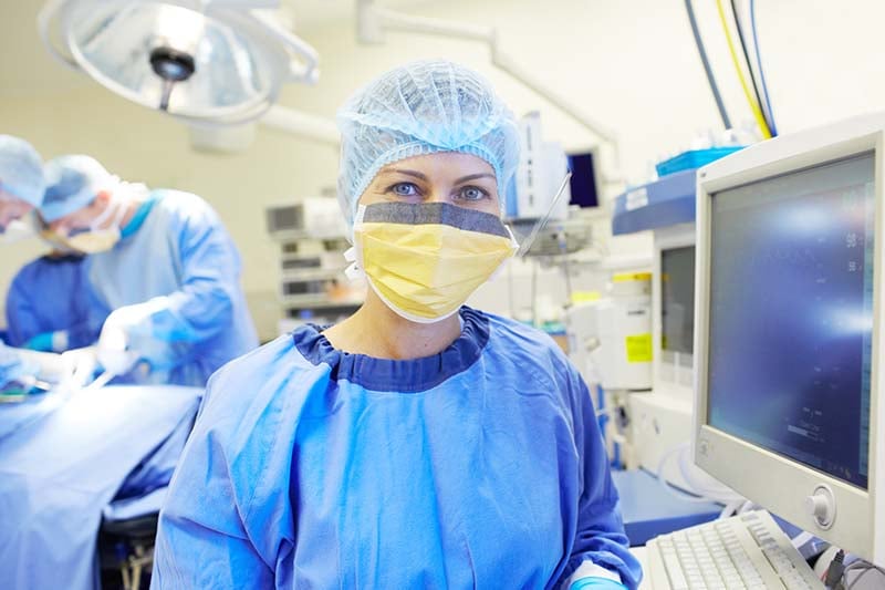A nurse is in the Operating Room standing near a keyboard and screen. They are wearing a surgical gown, hair covering and face mask. In the distance there is an obscured image of a surgical procedure taking place.