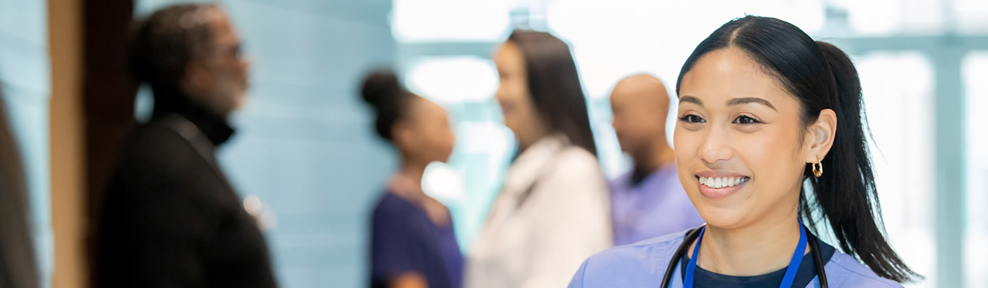A happy and smiling female nurse wearing blue scrubs is standing in a hospital corridor and taking with her co-workers.