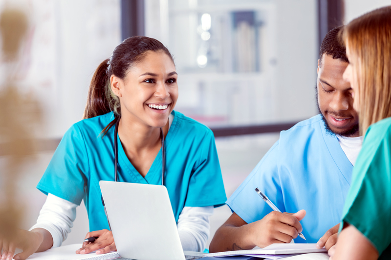 A group of three confident and smiling nursing students wearing scrub outfits are seated at a table with books and study materials in front of them, engaged in conversation. A male nursing student has his laptop open, and he is taking notes.