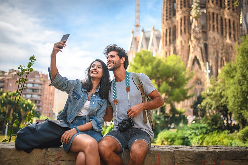 Male and female travelers are sitting on wall in public park near Sagrada Familia in Barcelona and taking a selfie on sunny summer day. Both are looking up at the phone and smiling.