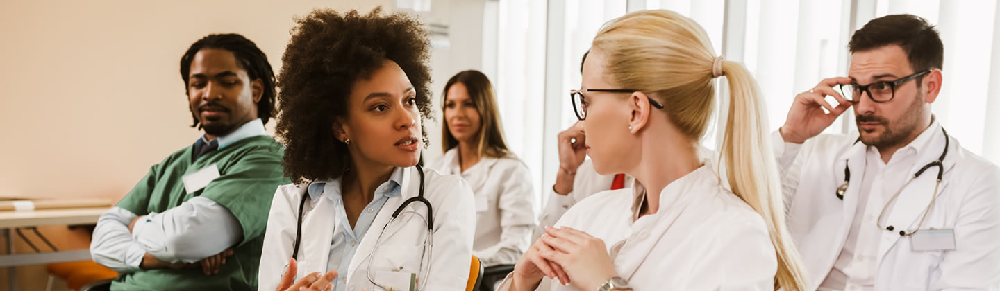 image depicts a group of healthcare professionals, likely a leadership team, engaged in a serious discussion. A woman with a curly afro speaks animatedly to a colleague, gesturing with her hands to emphasize her point, while others listen intently. The participants, wearing lab coats and scrubs, embody diversity and teamwork, key elements in resilient healthcare leadership.