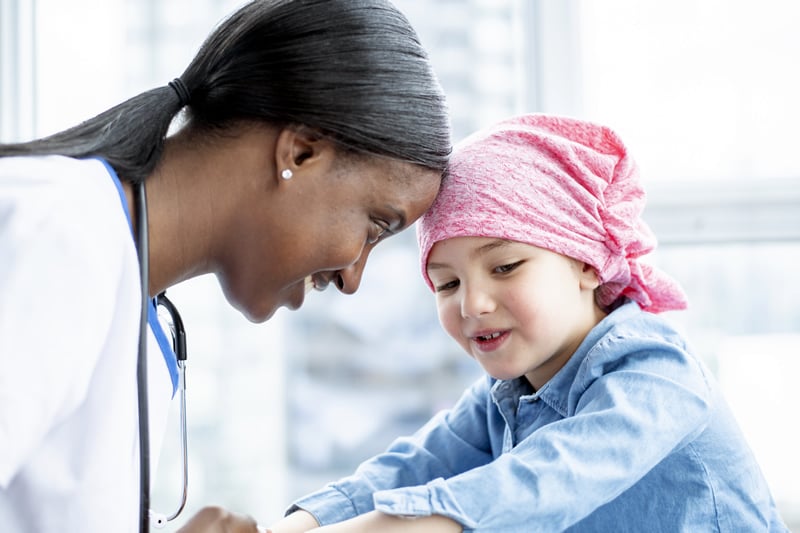 image captures a tender moment between a nurse and a young child wearing a pink headscarf, suggesting patient care in a pediatric oncology setting. The nurse, wearing a stethoscope, leans in with a smile to interact closely with the child, who looks up trustingly. This scene highlights the compassionate aspect of nursing leadership and the emotional resilience required in such sensitive healthcare environments.