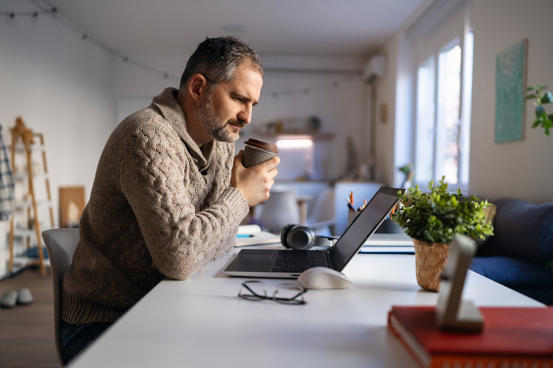 A middle-aged male nurse is at home, seated at a desk and looking at his laptop while studying online for his certification exam. He is wearing a light brown sweater and drinking a cup of coffee.