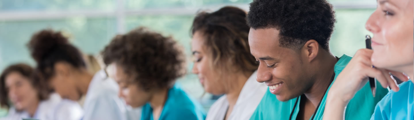 A group of diverse nurses are seated at a long conference table attending a continuing education course. Several nurses are taking notes, while others are looking forward and listening intently.
