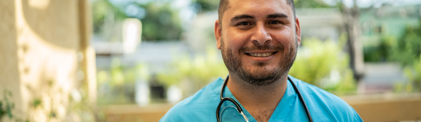 A confident male nurse is wearing blue scrubs and standing in an atrium area of a hospital. He is smiling and looking straight ahead.