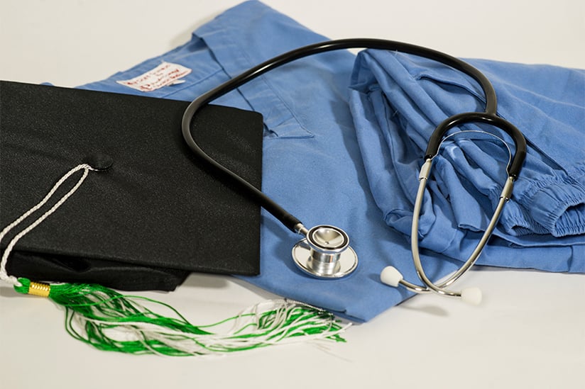 Image displays nursing educational achievements and tools: a dark academic cap with a green and white tassel rests atop a blue nurse's scrub top, accompanied by a stethoscope. This composition represents the completion of advanced nursing education and the readiness to apply the knowledge in a clinical setting.
