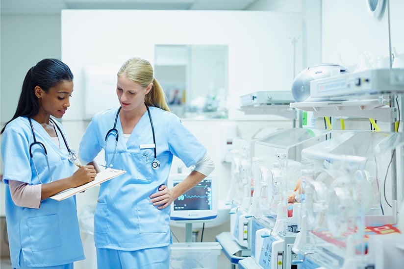 Two female nurses wearing light blue scrubs are standing in a neo-natal intensive care unit and looking at a clipboard while discussing a change of shift report. There are incubators and monitoring equipment lined up against a wall.