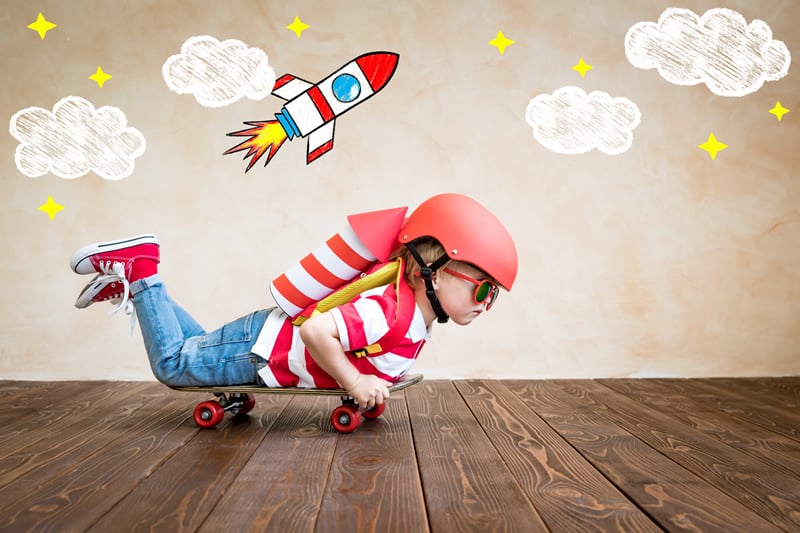 A young child is lying on a skateboard. They are wearing a red and white striped shirt and jeans, and there is a toy rocket strapped to their back and a helmet on their head. There is a drawing of clouds and a rocket blasting off above them. 