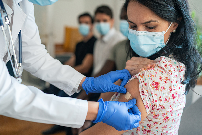 A nurse in a white lab coat and blue gloves is putting an adhesive bandage on the arm of a woman who is seated in a travel health clinic. She has just received a vaccination. There are several other people seated nearby waiting for their turn to be vaccinated.