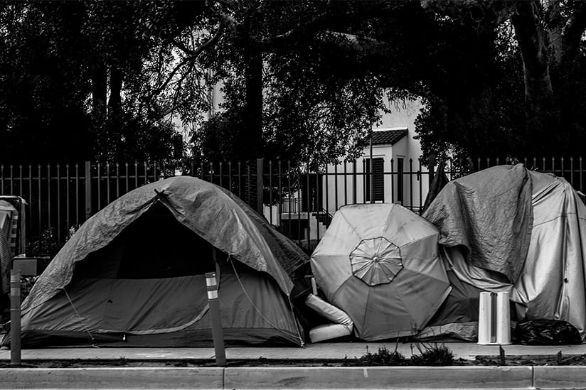 A black and white image of a row of tents and umbrellas set up on a city sidewalk. There are various blankets and possessions seen around the tents.