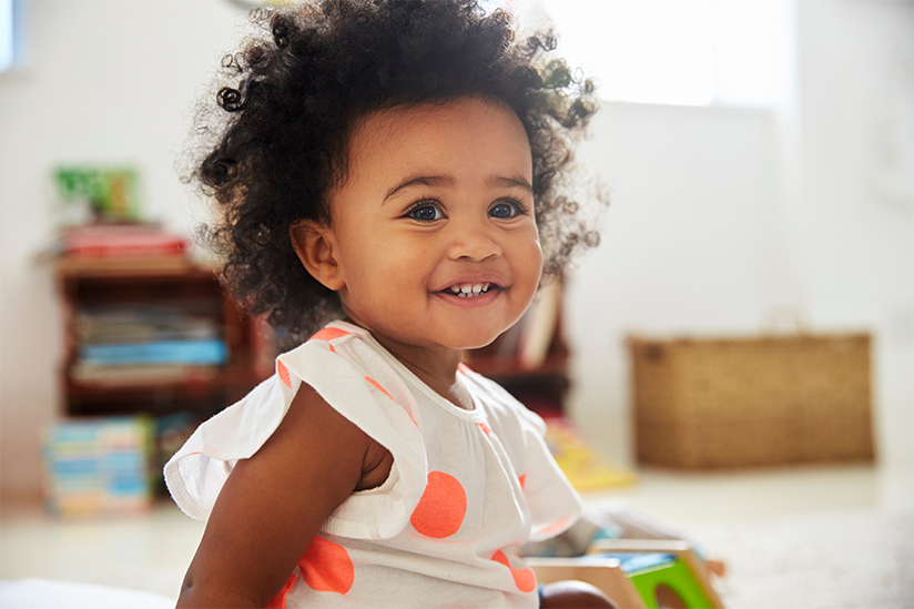 A beautiful baby with curly dark hair is wearing a white top with orange polka dots and a happy smile, and is seated on the floor with a toy. 