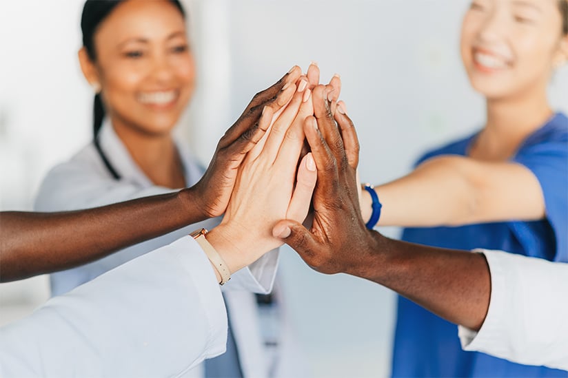 A group of nurses have extended their arms and placed their hands together, palms touching, in a circle signifying unity and teamwork. Only the forearms and hands are clearly visible.